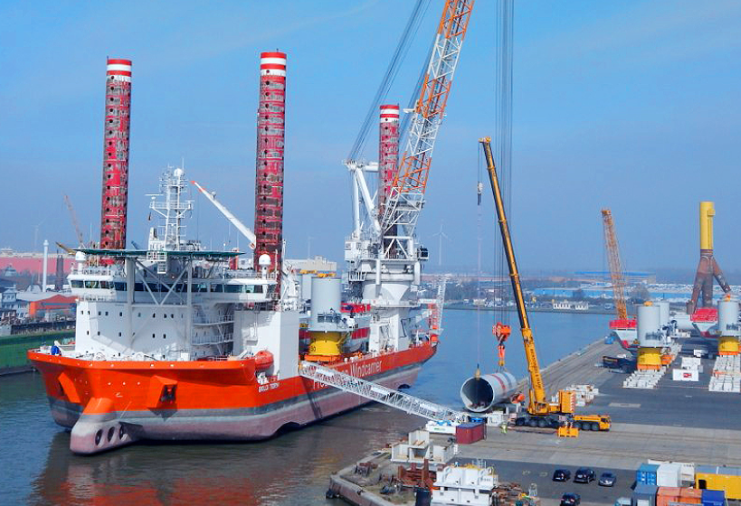 Spezialschiff mit roten Aufbauten im Hafen, Kräne bewegen Bauteile an Land.