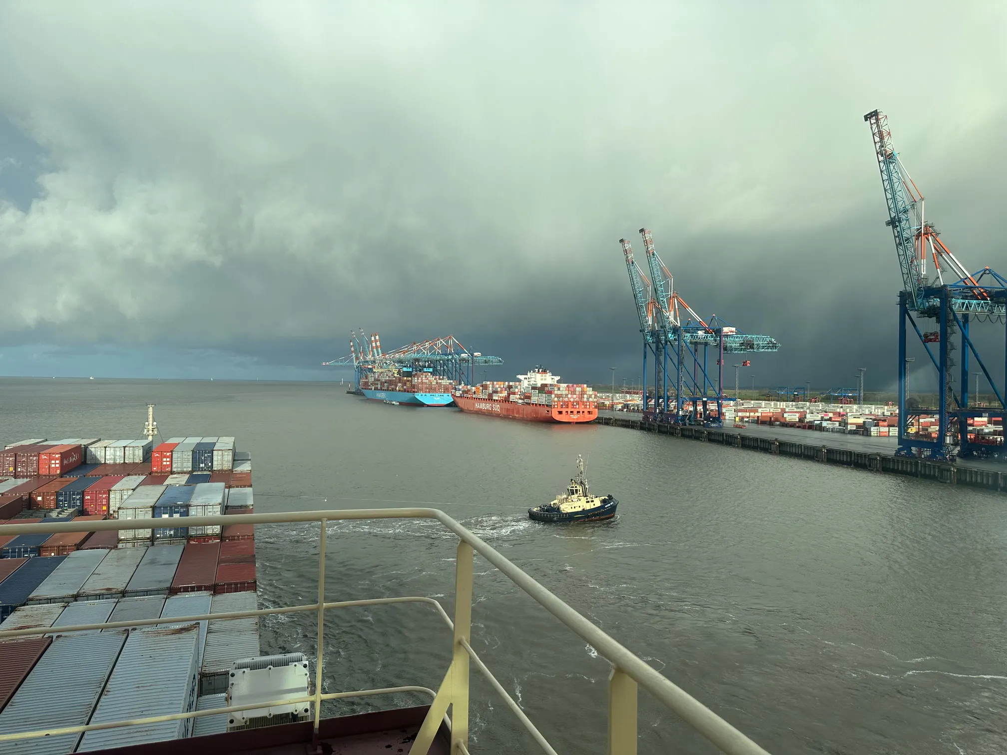 Containerhafen mit Containerschiffen und Hafenkränen, dunkle Wolken im Hintergrund.