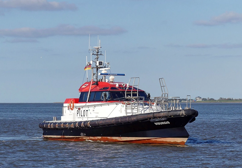 Das Bild zeigt ein modernes Lotsenboot auf einer Wasserfläche. Es ist weiß mit einem roten Dach, hat die Aufschrift "PILOT" und ist mit Sicherheitsausrüstung sowie Antennen ausgestattet. Im Hintergrund ist die Küste unter einem blauen Himmel zu sehen.